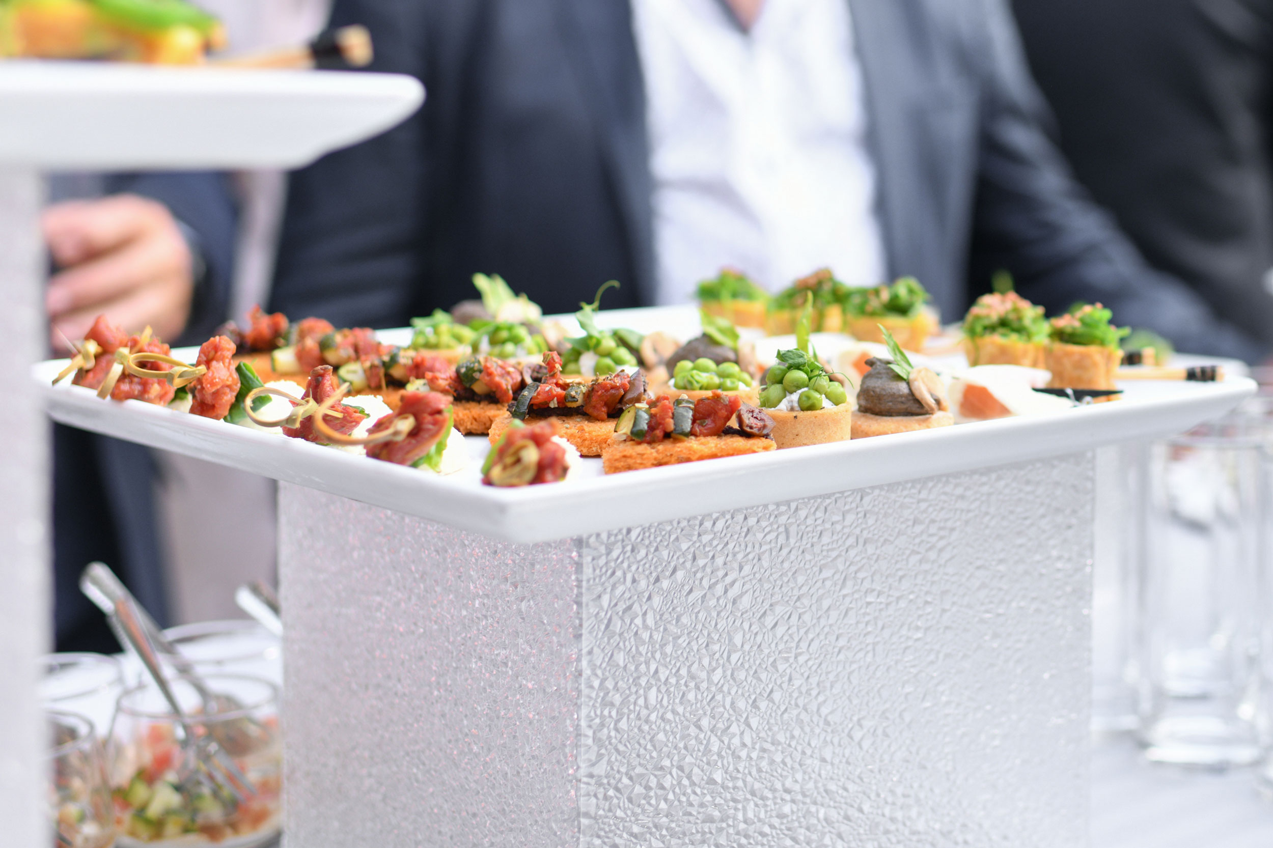 A close-up shot of a white platter filled with assorted appetisers, including small bite-sized pieces topped with various ingredients like diced vegetables, greens, and sauces. The platter is placed on a textured white stand. In the background, people in formal attire are seen, slightly out of focus. Some jars with utensils are visible at the bottom left corner.